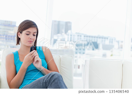 Adult woman sitting on white sofa beside glass windows, holding smartphone while gazing at skyline Adult woman sitting on white sofa beside glass windows, holding smartphone while gazing at skyline 135406540
