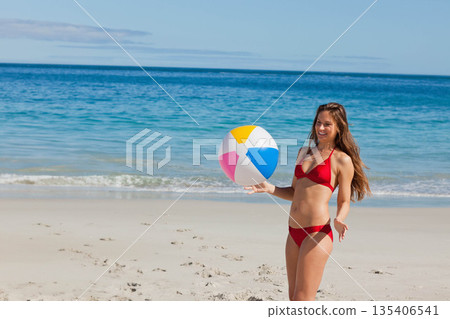 Woman standing on shore holding multicolored inflatable beach ball wearing red swimsuit, copy space 135406541