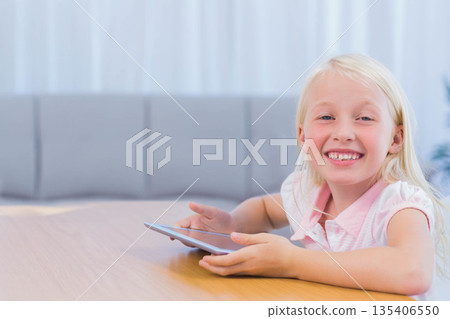 Female child sitting at light wood table inside home holding tablet and smiling, copy space Female child sitting at light wood table inside home holding tablet and smiling, copy space 135406550