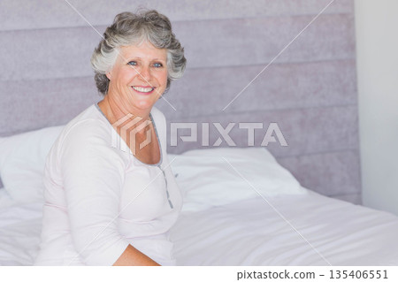 Senior woman sitting on bed smiling in bedroom with white linens and headboard, copy space 135406551