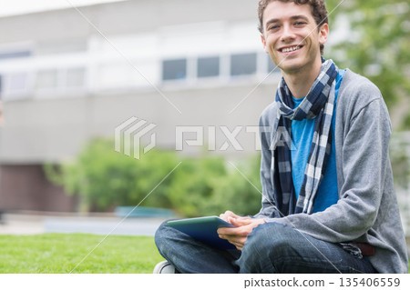 Adult male student sitting cross-legged on campus lawn holding tablet and smiling, copy space Adult male student sitting cross-legged on campus lawn holding tablet and smiling, copy space 135406559