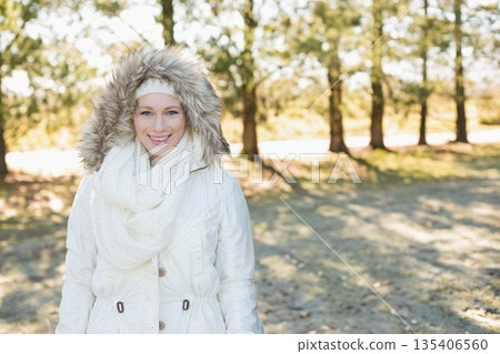 Woman standing in sunlit pine grove wearing white parka with faux fur trim, scarf and headband Woman standing in sunlit pine grove wearing white parka with faux fur trim, scarf and headband 135406560