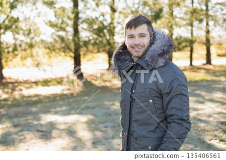 Man standing wearing winter coat under sunlit pine grove clearing, breathing chilly air, copy space Man standing wearing winter coat under sunlit pine grove clearing, breathing chilly air, copy space 135406561