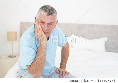 Senior man sitting on bed edge in bright bedroom holding head in hand beside nightstand lamp Senior man sitting on bed edge in bright bedroom holding head in hand beside nightstand lamp 135406571