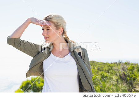 Woman hiking coastal hillside shading eyes and carrying backpack while wearing olive green jacket 135406579