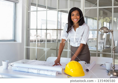 African American woman leaning forward reviewing blueprints on desk in office with yellow hard hat 135406580