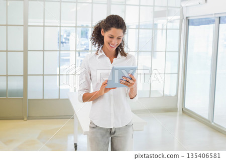 Woman in white shirt and gray jeans holding tablet and smiling while standing in modern office 135406581