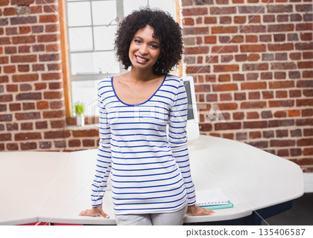 African American woman leaning on white desk in brick office with computer and window smiling African American woman leaning on white desk in brick office with computer and window smiling 135406587