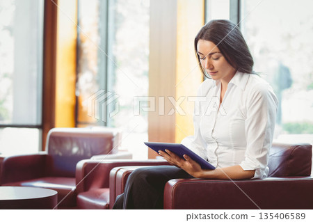 Woman holding tablet and reading while sitting in corporate lounge leather armchair, copy space 135406589