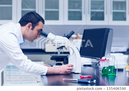 Asian male scientist leaning forward examining sample under microscope at lab bench with beakers 135406590