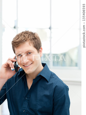 Man smiling and talking on smartphone while standing in office in front of large bright windows 135406593