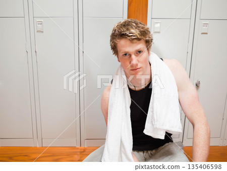 Fair-complexioned man sitting on bench by lockers in locker room holding white towel, copy space Fair-complexioned man sitting on bench by lockers in locker room holding white towel, copy space 135406598