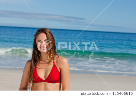 Woman standing on sandy beach at water's edge wearing red bikini top near rolling ocean waves Woman standing on sandy beach at water's edge wearing red bikini top near rolling ocean waves 135406610