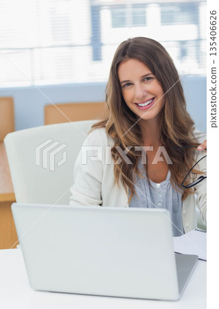Female professional holding eyeglasses and typing on silver laptop with notebook at office desk 135406626