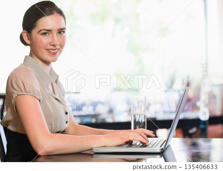 Woman sitting at wooden table in bright cafe working on laptop with water glass, copy space 135406633