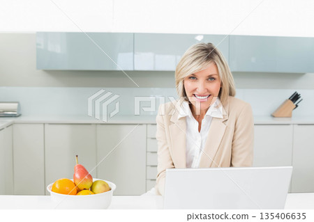 Middle-aged woman wearing blazer working on laptop at kitchen counter with fruit bowl, copy space Middle-aged woman wearing blazer working on laptop at kitchen counter with fruit bowl, copy space 135406635