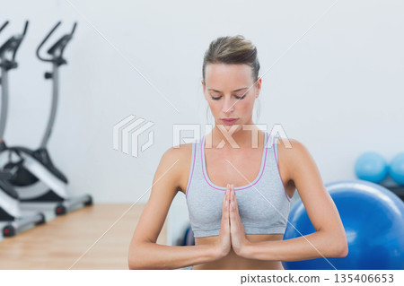 Woman practicing meditation pose in gym studio between elliptical machines and blue stability balls 135406653