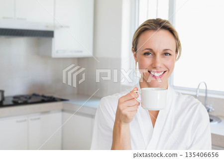 Woman smiling and holding white ceramic mug near mouth in kitchen with gas stove, copy space Woman smiling and holding white ceramic mug near mouth in kitchen with gas stove, copy space 135406656