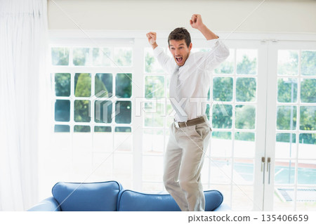 Man in his 30s jumping on blue couch wearing white shirt before grid windows showing pool Man in his 30s jumping on blue couch wearing white shirt before grid windows showing pool 135406659