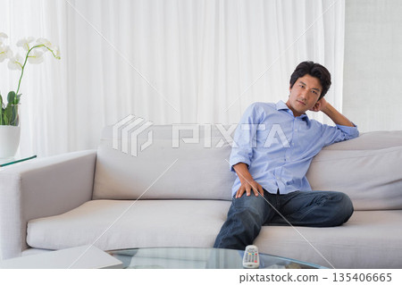 Japanese man sitting on gray sofa in bright living room with orchid and remote, copy space Japanese man sitting on gray sofa in bright living room with orchid and remote, copy space 135406665