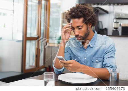 African American man sitting in denim shirt at cafe holding smartphone near plate and water glasses African American man sitting in denim shirt at cafe holding smartphone near plate and water glasses 135406673