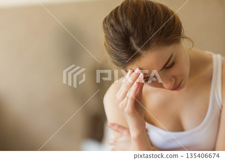 Woman sitting on beige couch in living room, wearing white tank top, touching forehead, copy space Woman sitting on beige couch in living room, wearing white tank top, touching forehead, copy space 135406674