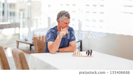Senior man sitting at sunroom table studying small wooden chessboard and chess pieces Senior man sitting at sunroom table studying small wooden chessboard and chess pieces 135406686