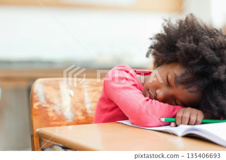 African American girl resting head on desk and chair holding green pencil over workbook at school African American girl resting head on desk and chair holding green pencil over workbook at school 135406693