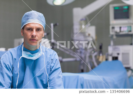 Male surgeon in blue scrubs standing in operating room near table under surgical lights, copy space Male surgeon in blue scrubs standing in operating room near table under surgical lights, copy space 135406696