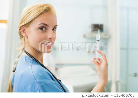 Female nurse wearing stethoscope holding and inspecting syringe beside bed in clinic, copy space 135406705