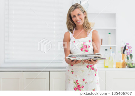 Mid adult woman standing in kitchen wearing floral apron holding newspapers with oils and orchid Mid adult woman standing in kitchen wearing floral apron holding newspapers with oils and orchid 135406707