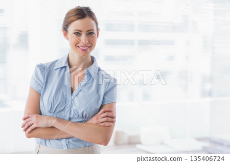 Mid-adult woman crossing arms wearing office attire in office with window blinds and desk 135406724