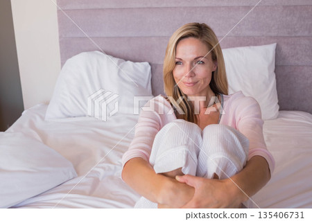 Woman sitting on bed hugging knees in bedroom with white linens and pink headboard Woman sitting on bed hugging knees in bedroom with white linens and pink headboard 135406731