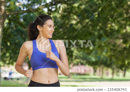 Female jogger running on grassy park path near bench wearing blue sports bra and black leggings 135406741
