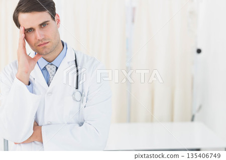 Male doctor standing pensively in examination room wearing lab coat beside stethoscope, copy space 135406749