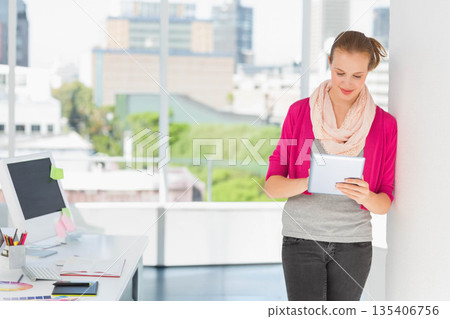 Woman reading tablet device at desk by office windows with computer monitor, copy space Woman reading tablet device at desk by office windows with computer monitor, copy space 135406756