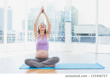 Mid adult pregnant woman practicing prenatal yoga on blue mat in bright studio with city skyline 135406757