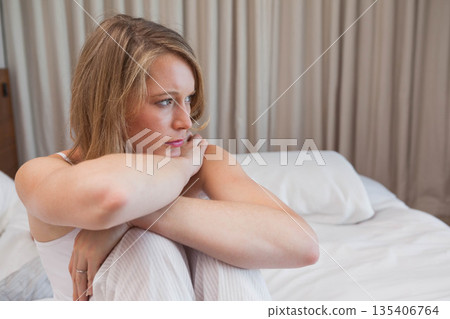 Woman sitting on bed in bedroom hugging knees and gazing right while wearing striped pajama pants Woman sitting on bed in bedroom hugging knees and gazing right while wearing striped pajama pants 135406764