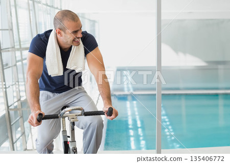 Man riding exercise bike at gym next to partition overlooking pool wearing white towel, copy space Man riding exercise bike at gym next to partition overlooking pool wearing white towel, copy space 135406772