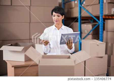 Woman holding tablet and wrapping cartons with bubble wrap at warehouse packing station 135406774
