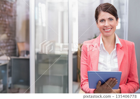 Mature woman wearing coral blazer holding tablet standing in lobby by glass partition, copy space 135406778