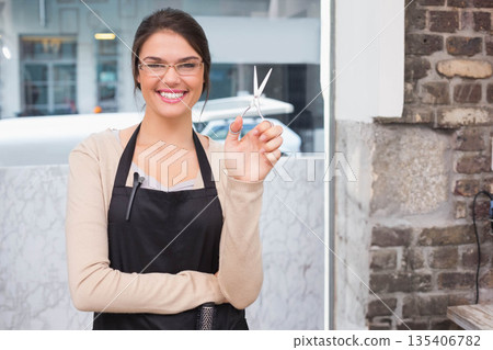 Female hairdresser wearing apron while holding shears and clip-on mic at marble counter in salon 135406782