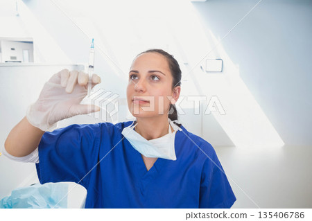 Woman medical professional in scrubs with mask around neck and gloves inspecting syringe in clinic 135406786