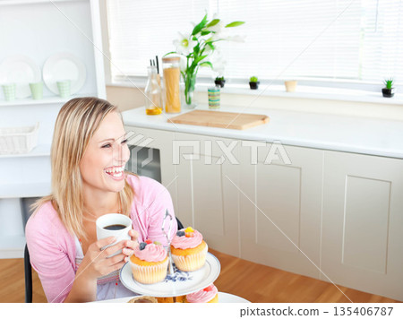 Smiling woman in pink cardigan in kitchen holding mug, gazing at frosted cupcake stand, copy space Smiling woman in pink cardigan in kitchen holding mug, gazing at frosted cupcake stand, copy space 135406787