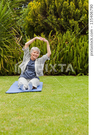 Senior woman sitting on blue yoga mat in backyard stretching arms overhead wearing gray sportswear 135406790
