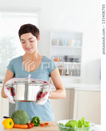 Middle-aged female wearing floral mitts holding pot and chopping vegetables in kitchen, copy space 135406793