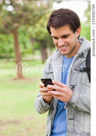 Man standing in park holding smartphone and smiling in denim jacket and backpack, copy space 135406799