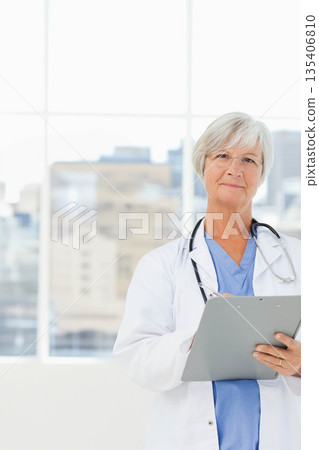 Senior doctor standing by window in clinic with stethoscope, reviewing clipboard, copy space 135406810
