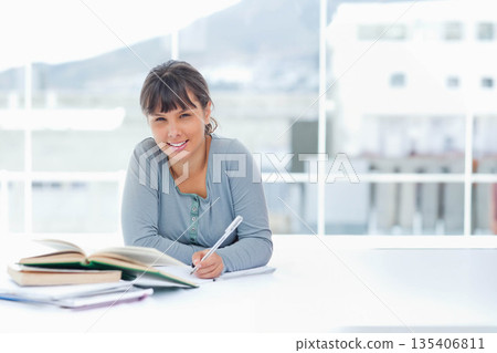 Female student sitting at study table by large windows with open textbooks, writing in notebook 135406811