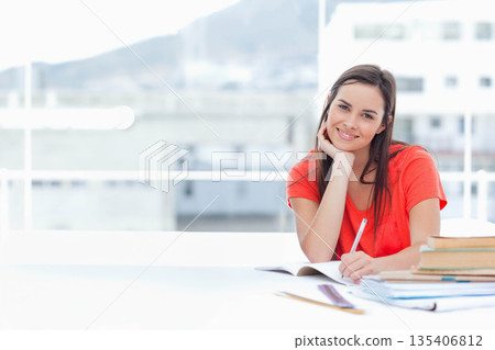 Woman writing in notebook at desk by railing bars wearing red t-shirt beside textbooks, copy space Woman writing in notebook at desk by railing bars wearing red t-shirt beside textbooks, copy space 135406812